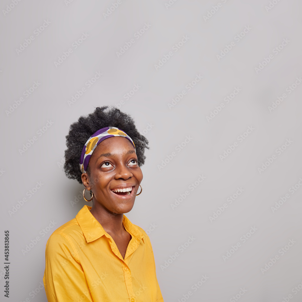Vertical shot of dark skinned curly woman focused above smiles broadly has happy mood reads promo text overhead wears headband and yellow shirt poses against grey background blank copy space Vertical shot of dark skinned curly woman focused above smiles broadly has happy mood reads promo text overhead wears headband and yellow shirt poses against grey background blank copy space
