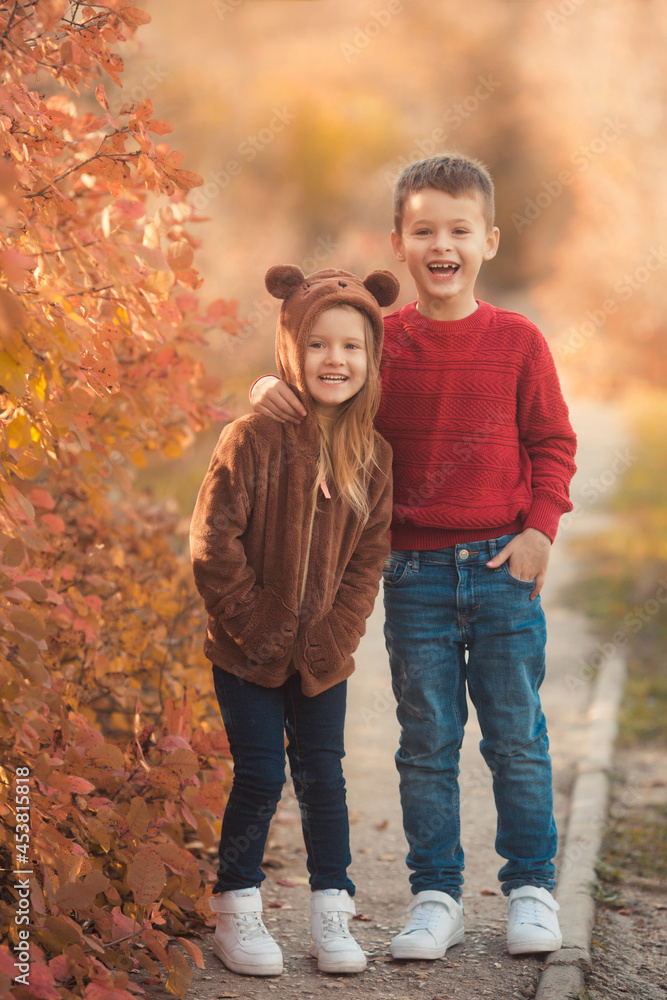 Happy friends hugging on a walk in the fall park. Portrait of a caucasian sister and brother in beautiful outfits on a sunny autumn day in forest. Family lifestyle concept.