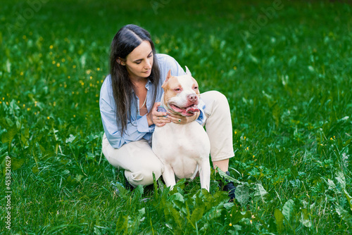 Brunette female in casual clothes patting bicolor white-brown active american pitbull terrier dog on grass in city park. Walking and training dog outside. Happy animal with tongue out looking away photo