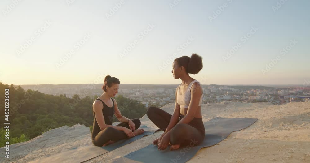 Mindful peaceful diverse women with a calm look chatting with each other and meditates outdoors, while sitting at the yoga mat. Females trying to relax after long hours of working Mindful peaceful diverse women with a calm look chatting with each other and meditates outdoors, while sitting at the yoga mat. Females trying to relax after long hours of working