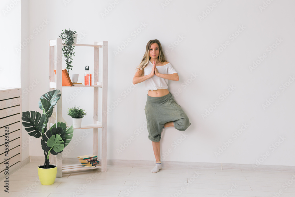 Woman practise yoga performs. Tree pose standing on mat inside of cozy room with plants and greenery. Healthy lifestyle concept Woman practise yoga performs. Tree pose standing on mat inside of cozy room with plants and greenery. Healthy lifestyle concept