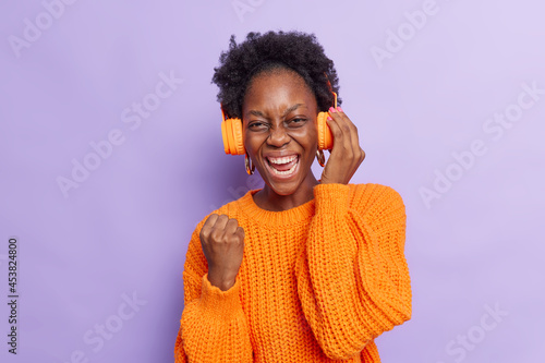 Favorite music fun and pleasure concept. Joyful millennial girl with Afro hair clenches fist makes yes gesture enjoys good sound in modern headphones listens popular song wears knitted sweater