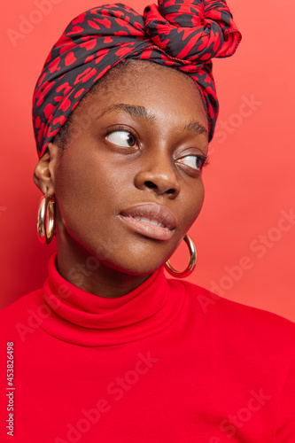 Close up portrait of serious dark skinned ethnic lady looks with calm face expression at someone wears casual turtleneck kerchief on head poses against vivid red background being deep in thoughts