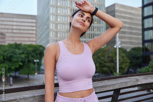 Tired sportswoman wipes forehead after hard fitness training wears cropped top smartwatch looks thoughtfully forward poses at bridge against tall cityscrapers view being in good physical shape © wayhome.studio 