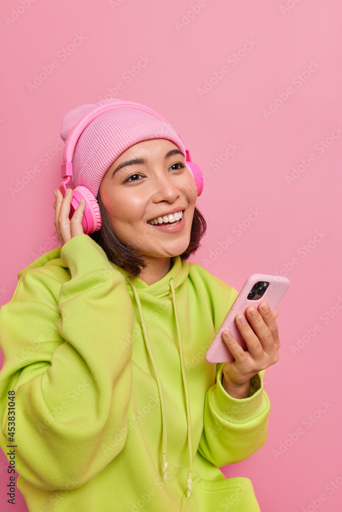 Vertical shot of happy Asian teenage girl holds mobile phone in hand enjoys listening music from playlist has pleasant smiles on face dressed casually. Cheerful female student listens audio book Vertical shot of happy Asian teenage girl holds mobile phone in hand enjoys listening music from playlist has pleasant smiles on face dressed casually. Cheerful female student listens audio book