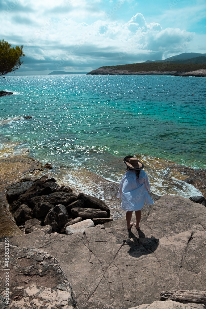 Mistery woman seen from behind wearing a white large shirt and a hat. Teal amazing adriatic sea stretching in the distance. Srebrna beach on vis island in croatia. Summer of 2021