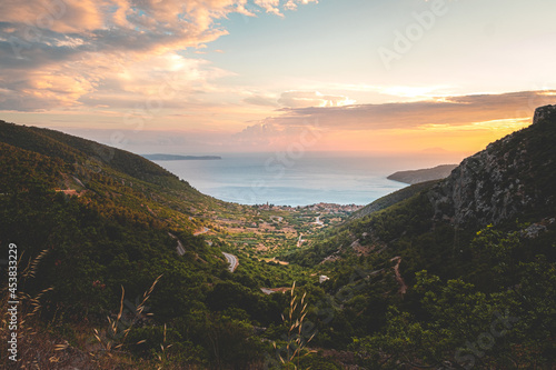 Amazing sunset on the Vis island in Croatia. Komiza city in the distance. Golden hour, orange sky and the beautiful blue adriatic sea. Green mountains surrounding the little picturesque town