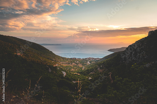 Amazing sunset on the Vis island in Croatia. Komiza city in the distance. Golden hour, orange sky and the beautiful blue adriatic sea. Green mountains surrounding the little picturesque town