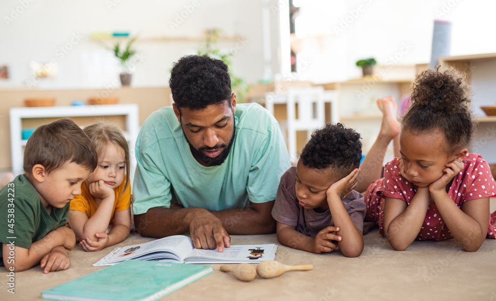 Group of small nursery school children with man teacher on floor indoors in classroom, reading book. Group of small nursery school children with man teacher on floor indoors in classroom, reading book.