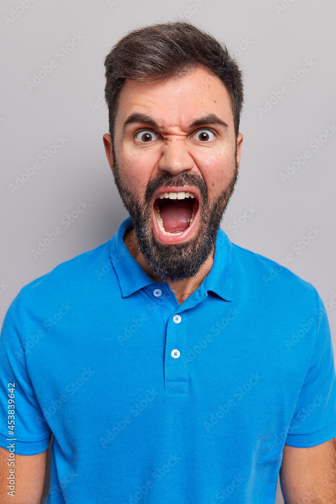 Outraged pissed bearded European man shouts angrily keeps mouth widely opened expresses negative emotions screams furious wears casual blue t shirt isolated over grey background shouts from scorn Outraged pissed bearded European man shouts angrily keeps mouth widely opened expresses negative emotions screams furious wears casual blue t shirt isolated over grey background shouts from scorn