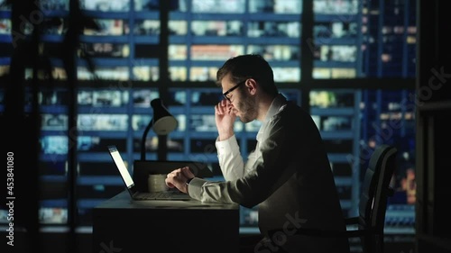 Tired young man working on a laptop late night in the office. Sleepy Businessman sitting at desk in dark office. Tired and stressed businessman in glasses works on a laptop of the night city office