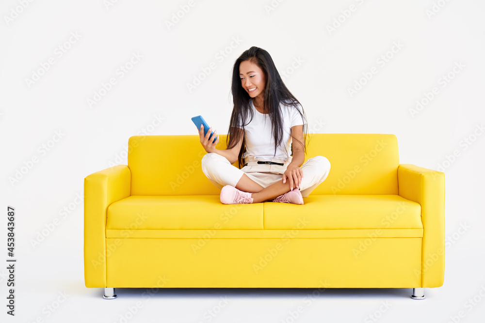 Smiling asian woman holding smartphone browses internet and scrolls news sitting on yellow couch Smiling asian woman holding smartphone browses internet and scrolls news sitting on yellow couch