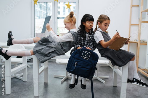 Wild little schoolgirls sitting on the desk, having fun. Leaning on each other.