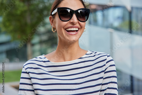 Outdoor shot of glad dark haired woman smiles broadly shows white teeth wears trendy sunglasses striped jumper poses against blurred background strolls in open air. People and lifestyle concept