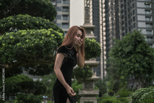 Portrait of beautiful young asian woman in casual black dress standing in the park and laughing. Covers her mouth with her hand. Long hair