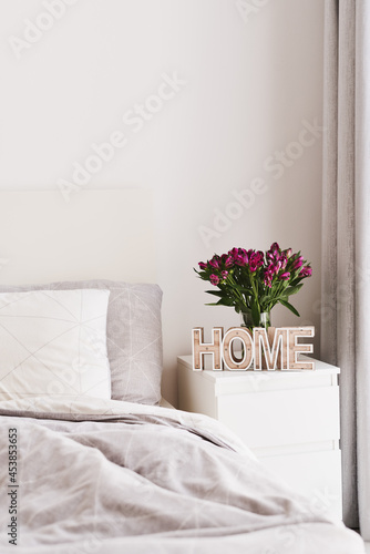 Bedroom decor: white bed with grey bedding and two pillows, white bedside table with wooden "home" sign and fresh Alstroemeria flowers in a vase. Minimal scandinavian interior design