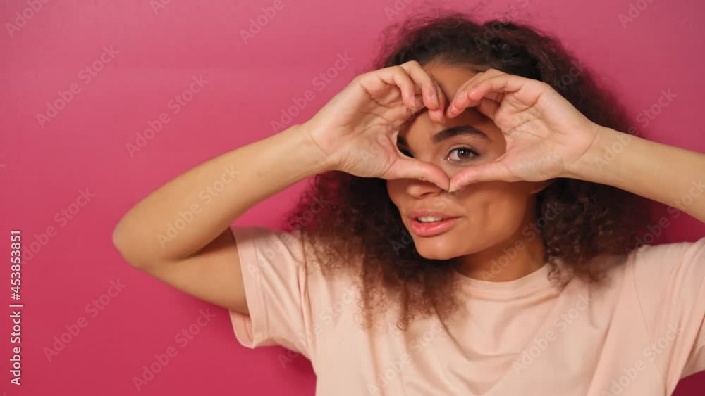 Peeps through the heart or love gesture beautiful young African American woman looking positively at camera wearing peachy t-shirt isolated on pink background. Beauty concept. 4kFootage. Peeps through the heart or love gesture beautiful young African American woman looking positively at camera wearing peachy t-shirt isolated on pink background. Beauty concept. 4kFootage.