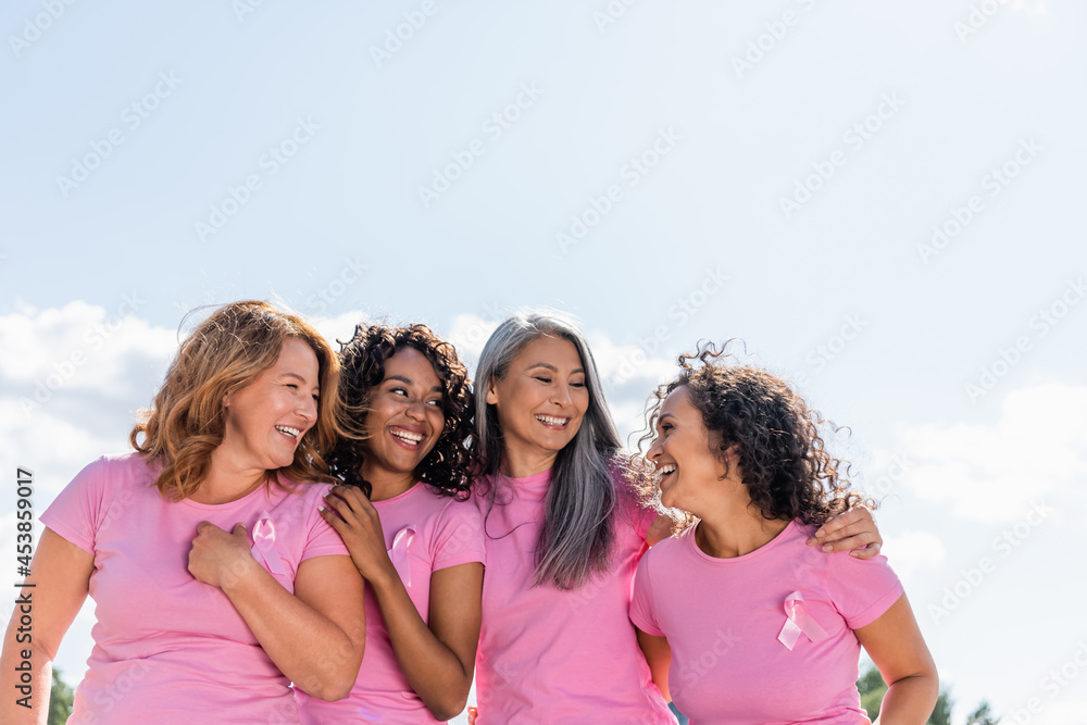 Cheerful multiethnic women with ribbons of breast cancer awareness hugging outdoors Cheerful multiethnic women with ribbons of breast cancer awareness hugging outdoors