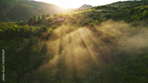 Sunrise over misty forest, warm rays of sunlight and distant cabin or farm house landscape