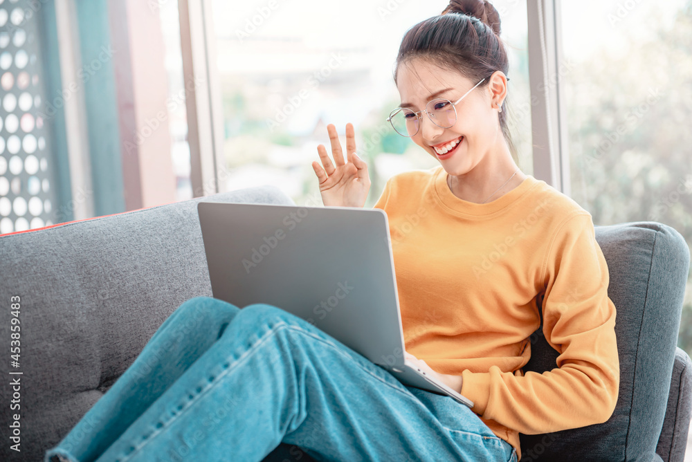 Naklejka premium Happy asian woman using videoconferencing with her friend via online app on laptop, waving hand and saying Hi
