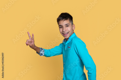 Young smiling teenage boy  gestures a sign  victory or pease against a yellow background.