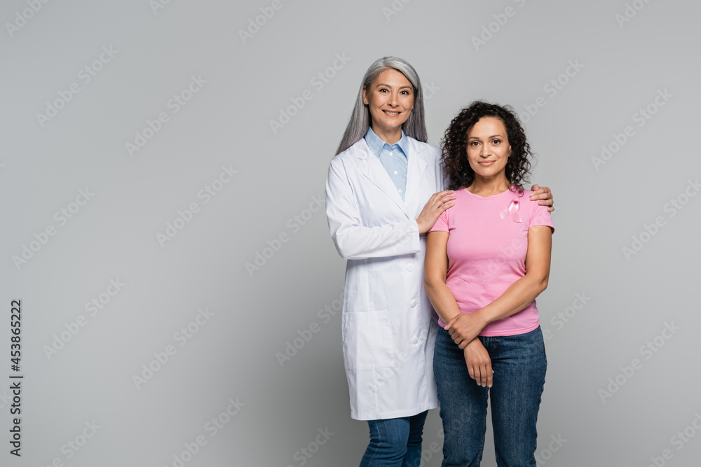 Smiling asian doctor in white coat hugging african american patient with ribbon of breast cancer awareness isolated on grey Smiling asian doctor in white coat hugging african american patient with ribbon of breast cancer awareness isolated on grey