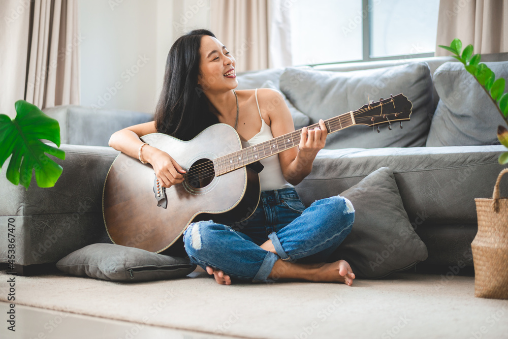 Asian woman playing music by guitar at home, young female guitarist musician lifestyle with acoustic art instrument sitting to play and sing a song making sound in hobby in the house room - obrazy, fototapety, plakaty 