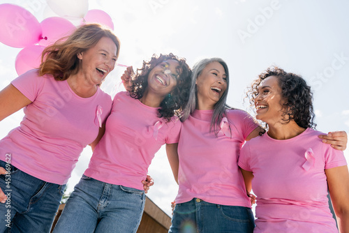 Low angle view of cheerful interracial women with pink ribbons and balloons hugging outdoors photo