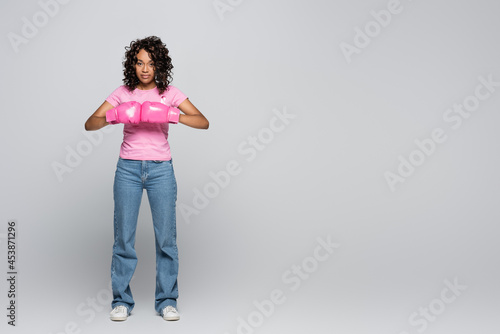 Young african american woman in t-shirt with ribbon and boxing gloves on grey background