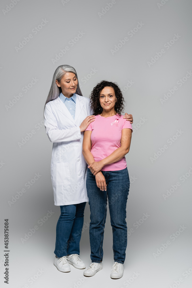 Smiling asian doctor hugging african american woman with ribbon of breast cancer awareness on grey background Smiling asian doctor hugging african american woman with ribbon of breast cancer awareness on grey background