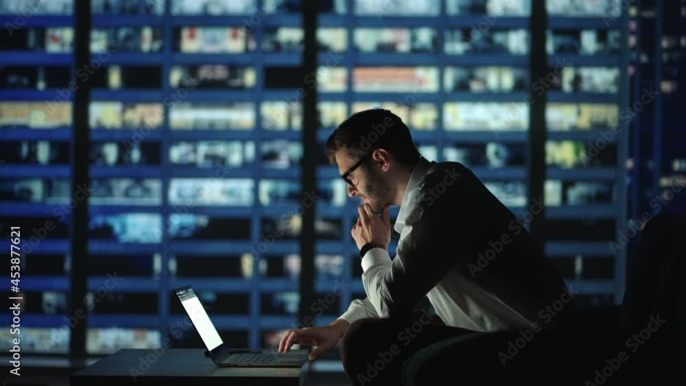Focused business man working on laptop computer making notes sitting at home office desk at night. Dark room city and Young adult male professional manager doing marketing research Focused business man working on laptop computer making notes sitting at home office desk at night. Dark room city and Young adult male professional manager doing marketing research