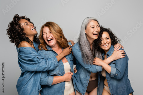 Happy multiethnic women in underwear and denim shirts embracing isolated on grey, body positive concept photo