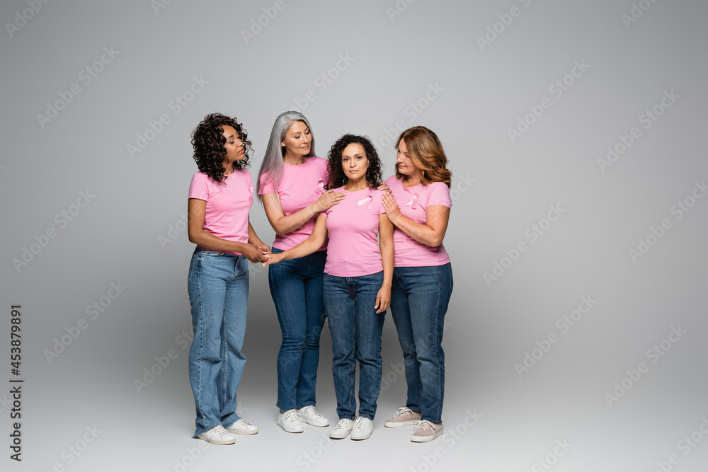 Multiethnic women hugging friend with pink ribbon on t-shirt on grey background Multiethnic women hugging friend with pink ribbon on t-shirt on grey background