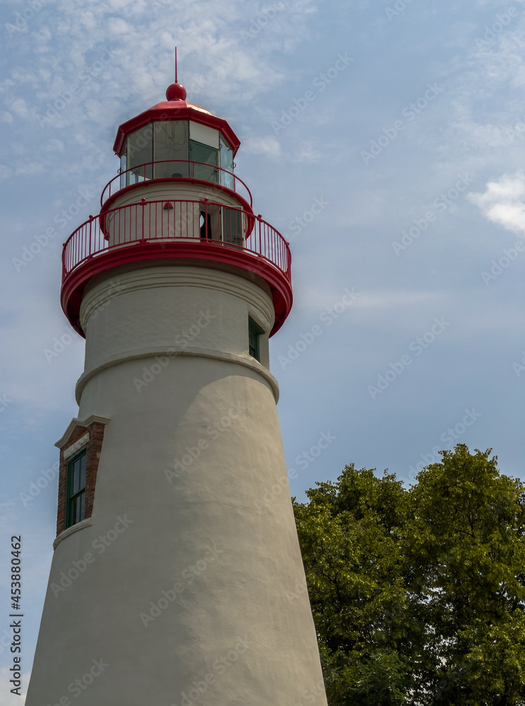 The red and white Marblehead Lighthouse with cloudy blue sky and green trees in background
