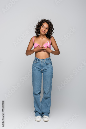 Smiling african american woman in bra with ribbon of breast cancer awareness on grey background