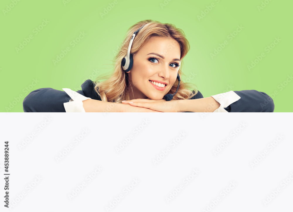 Call center. Customer support service female phone operator or sales agent in headset, grey confident suit standing behind mock up signboard with copy space area, isolated over light green color back. Call center. Customer support service female phone operator or sales agent in headset, grey confident suit standing behind mock up signboard with copy space area, isolated over light green color back.