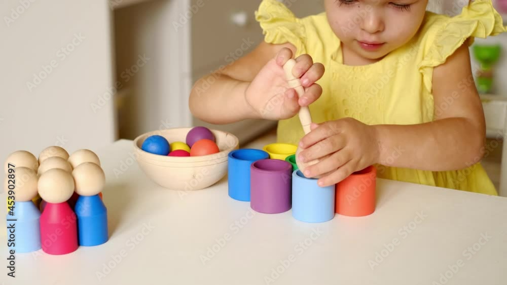 The child holds a wooden spoon with a ball in his hands, which he later throws into a cup of the same color. A girl learns colors by playing a sorter toy. Modern exciting games for preschool education The child holds a wooden spoon with a ball in his hands, which he later throws into a cup of the same color. A girl learns colors by playing a sorter toy. Modern exciting games for preschool education