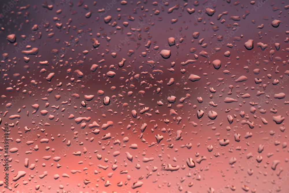 Szyba okienna pokryta od zewnątrz kroplami deszczu. Za oknem widać chmury zabarwione na czerwono światłem zachodzącego słońca. - obrazy, fototapety, plakaty 