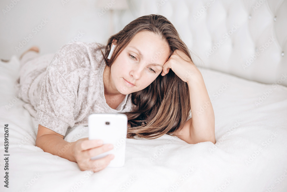 Beautiful brunette girl is lying on bed with wireless headphones and white smartphone in her hands and listening to music or audiobook. Beautiful brunette girl is lying on bed with wireless headphones and white smartphone in her hands and listening to music or audiobook.