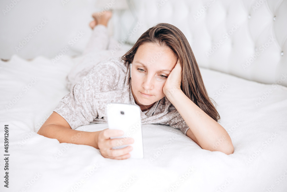 Beautiful brunette girl is lying on bed with wireless headphones and white smartphone in her hands and listening to music or audiobook.
