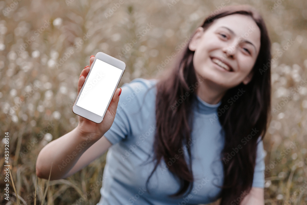Pretty brunette teenage girl with smartphone outdoors in park.  Mock-up concept. Smilling girl showing a blank smartphone screen to camera, for mobile ads and web services, copy space. Empty screen.