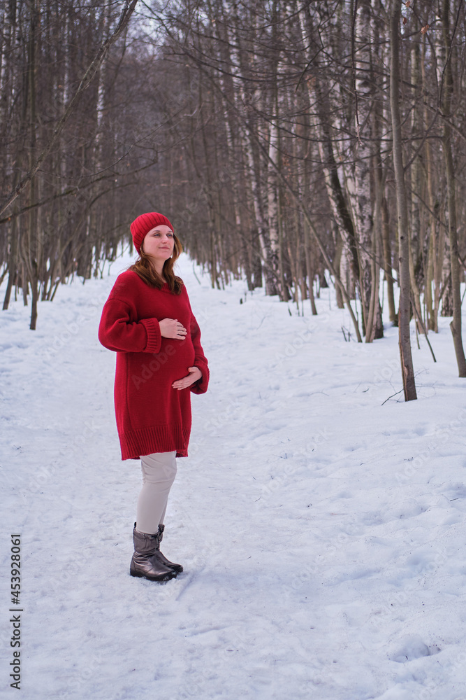 A happy pregnant woman in a knitted sweater and a hat walks in the park, a winter forest with trees in the snow