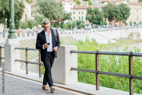 Urban businessman wearing jacket and shoulder bag. Handsome male model in his 45s. Young entrepreneur walking in the street and holding a mobile smartphone using app text sms message photo