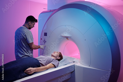 Young doctor pushing button on panel to start mri scan procedure while female patient lying on long table photo