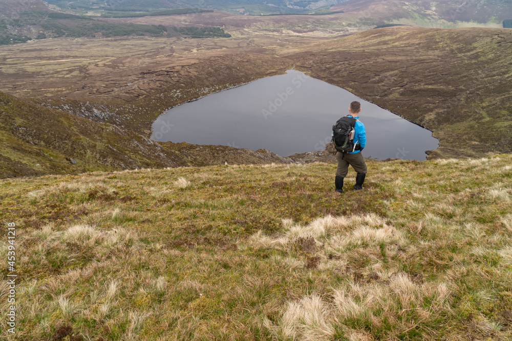 Hiker is standing on the top of the mountain with hands in the pockets. Back view.. Heart shaped Lake Ouler Tonelagee Mountain, Wicklow County, Ireland.