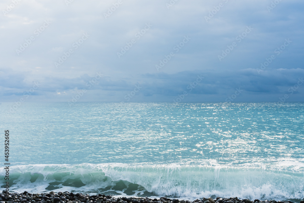 Fototapeta premium A wave of turquoise sea shining in the sun runs over black pebbles. The sea on the horizon merges with the blue sky.