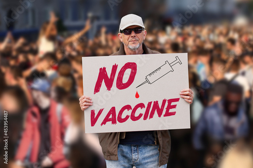 Displeased man with cap, blue jeans and sunglasses holding a NO COVID vaccine sign with crowd of people in background. Supporting anti-vaccination movement. Anti-vaxxer activist.  © Ole