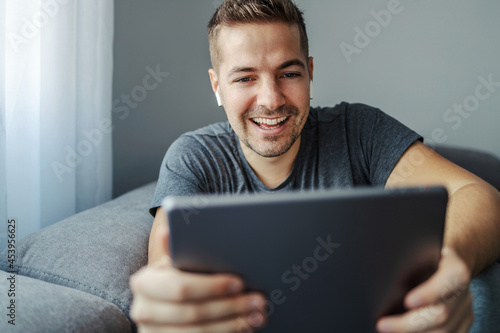 Online chat and internet messaging, phone call. A man with a beautiful smile discusses with friends and colleagues. He has white wireless headphones and uses a gray tablet that he holds in his hands