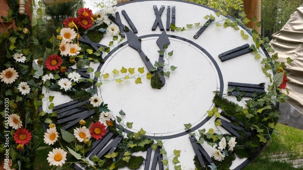 Large wall clock on the field in blooming flowers, showing the time 11:55, 12:00. time management concept, daylight saving time. Spring or wedding. Picnic for womens day