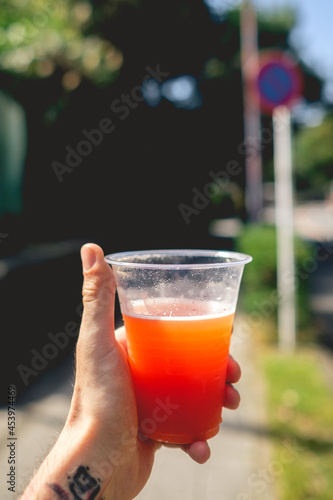 White hand with korean tattoo in the wrist holding a plastic cup with tomato beer under the sunlight